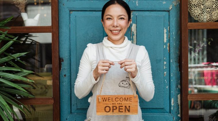 Cheerful ethnic female cafeteria owner in apron demonstrating cardboard signboard while standing near blue shabby door and windows after starting own business and looking at camera