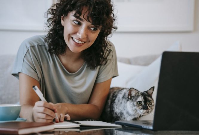 Crop young positive female smiling and taking notes in organizer while cat watching on netbook at table at home