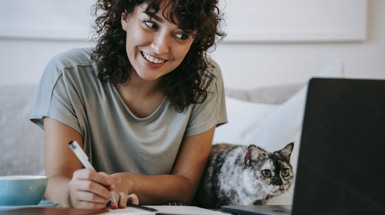 Crop young positive female smiling and taking notes in organizer while cat watching on netbook at table at home