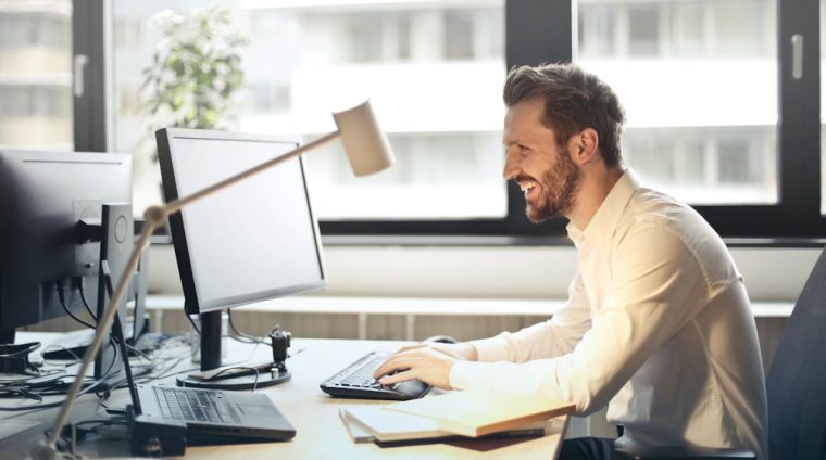 Man in White Dress Shirt Sitting on Black Rolling Chair While Facing Black Computer Set and Smiling